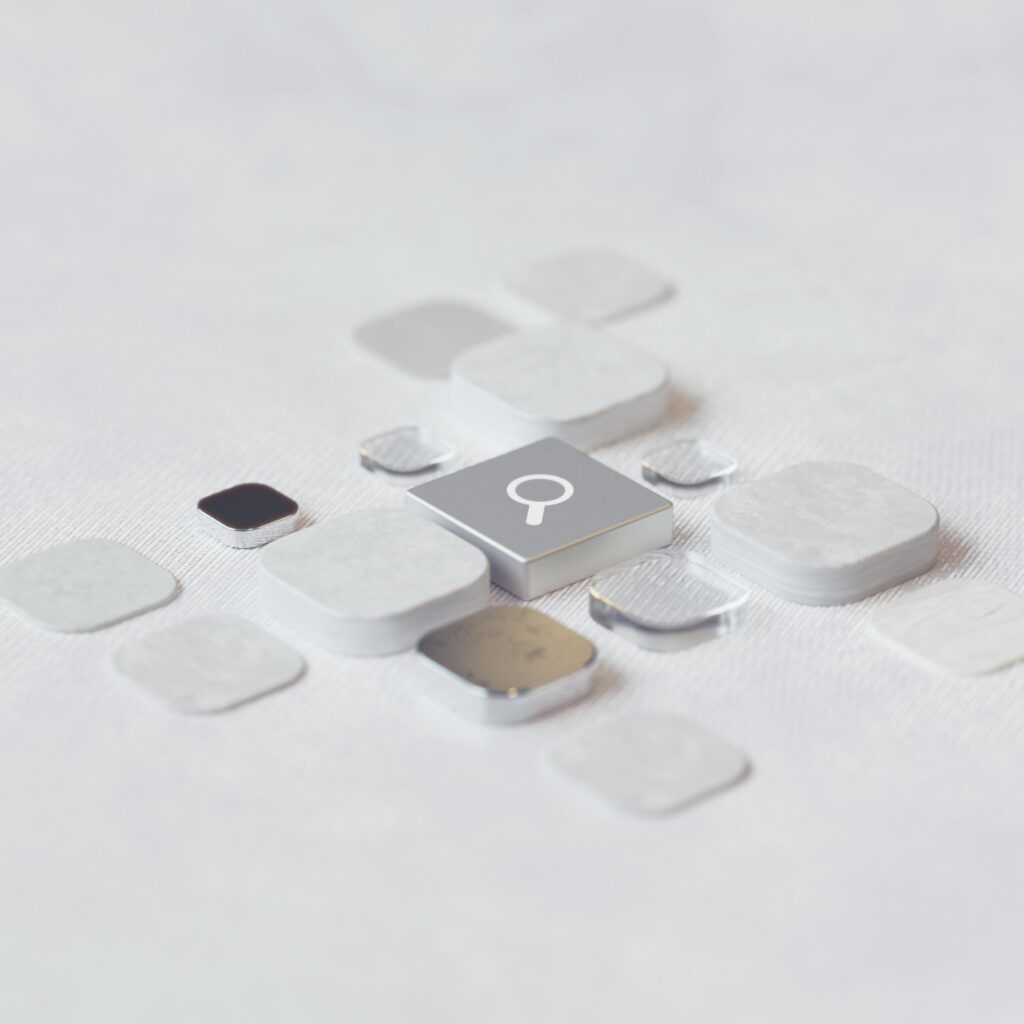 Abstract image featuring a magnifying glass icon on a square gray button, symbolizing search or discovery in AI, surrounded by scattered translucent and solid tiles in various shapes, set on a textured white surface.