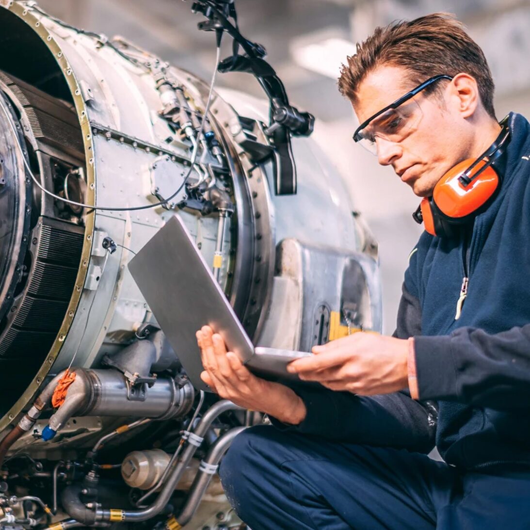 engine mechanic looking at laptop in front of large engine